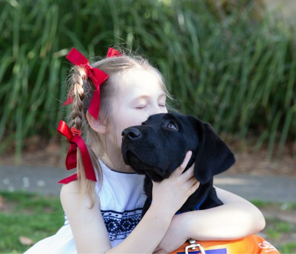 A young child kissing a black twelve week old black labrador puppy. the young child has its eyes closed and the puppy is looking to the left of the camera.